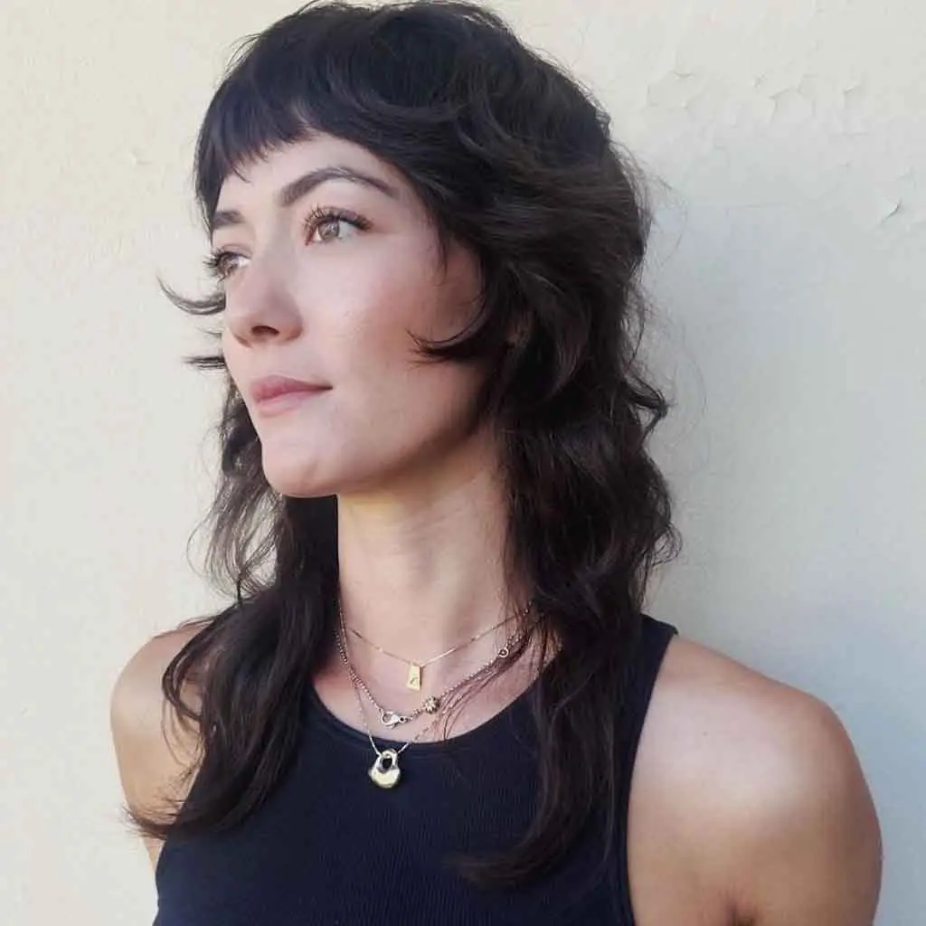 Woman displaying a medium raven black wavy mullet wolf cut with micro bangs, blending soft waves with an edgy touch