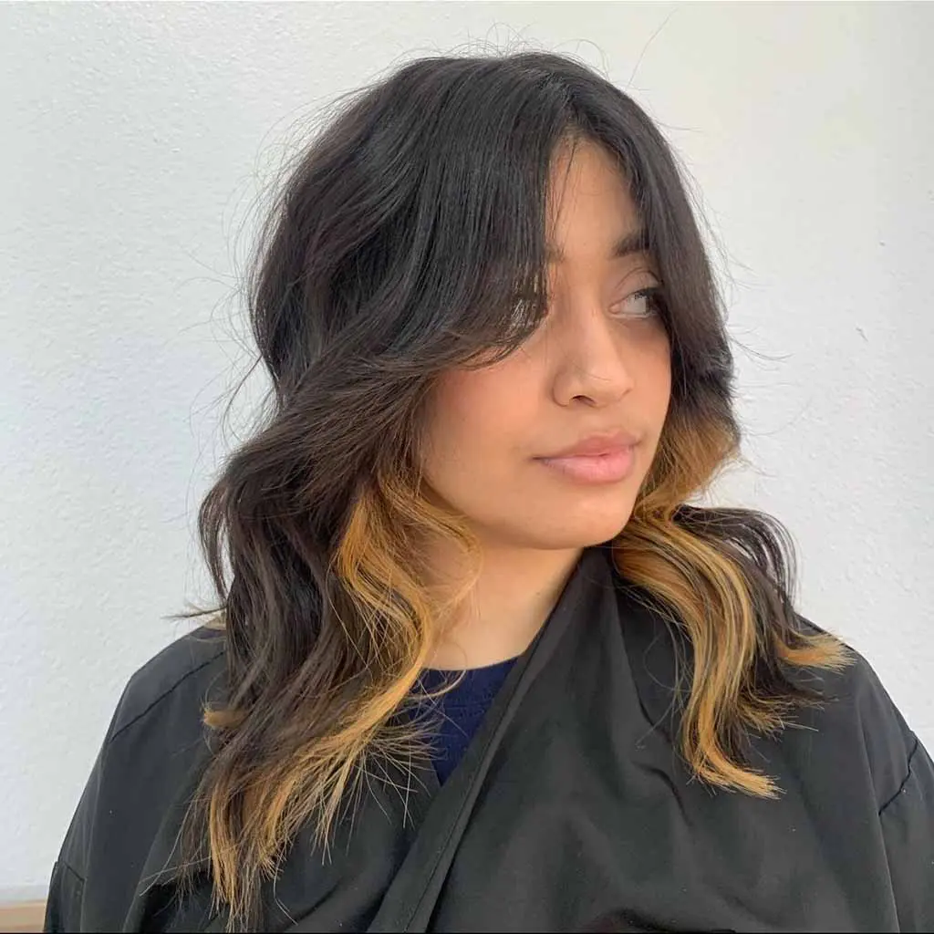 Woman showing off a medium wavy black plum wolf cut with curtain bangs and bronde chunky highlights, blending rich hues with dimension