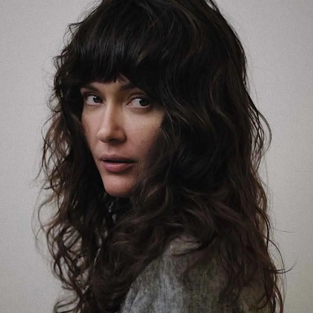 Woman flaunting a midnight black wavy and shaggy long wolf cut with bold blunt bangs, adding texture and movement