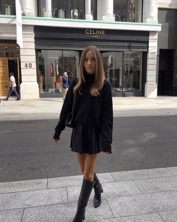 A woman in a black outfit including a sweater pleated skirt and knee high boots