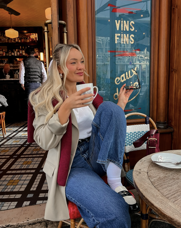 A woman sits at a cafe table holding a coffee cup and wearing a beige coat with a burgundy scarf. She has long blonde hair and is dressed in jeans and loafers