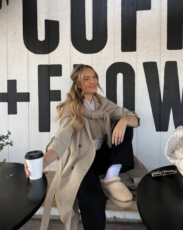 beige trench coat with a sweater draped over their shoulders black pants and cozy slippers