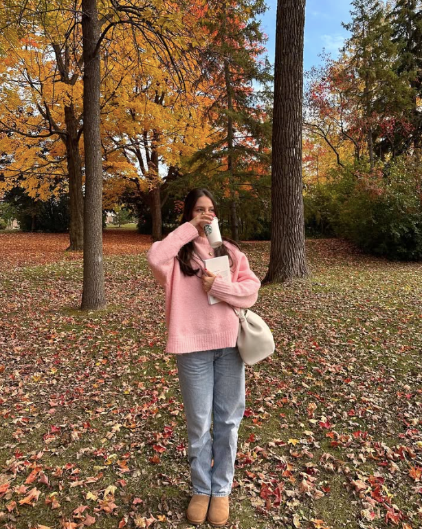 pink sweater stands in a park with vibrant autumn foliage sipping from a Starbucks cup. She holds a book and wears light blue jeans and brown boots