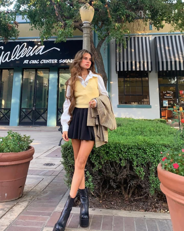 woman wearing a yellow vest over a white shirt a black skirt and black platform boots holding a plaid jacket