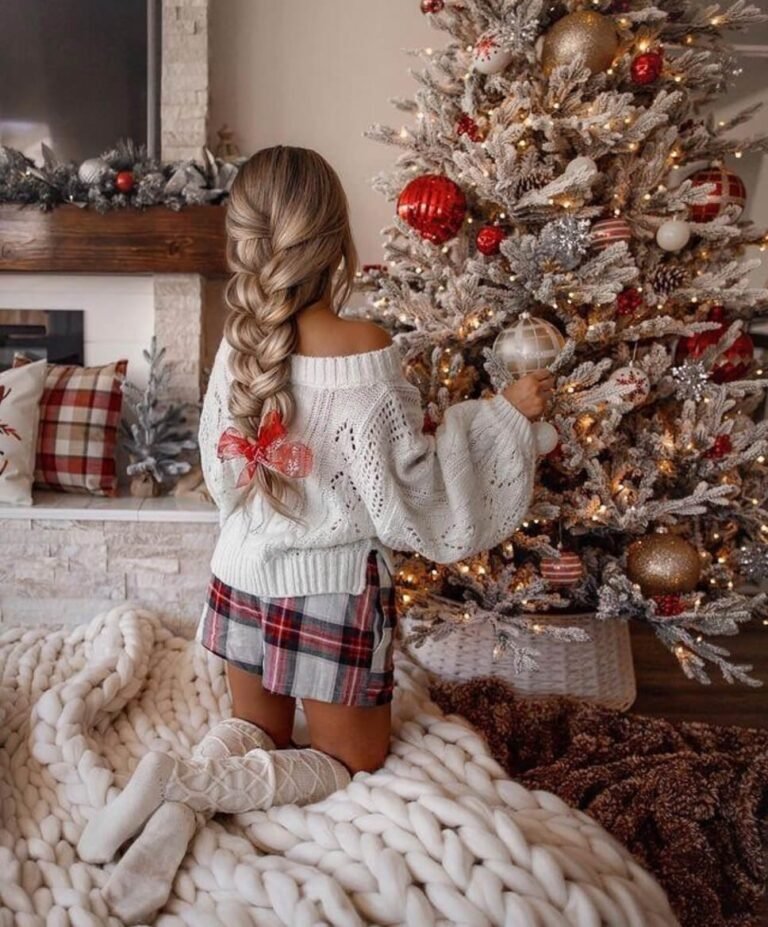 A woman with a long braid kneels on a chunky knit blanket, decorating a Christmas tree with red and gold ornaments.